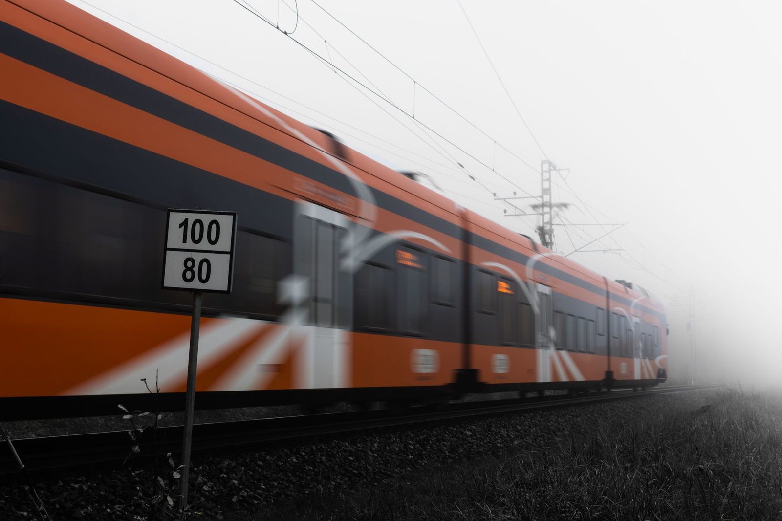 Train régional orange et gris en mouvement sur une voie électrifiée dans le brouillard, près d'un panneau de vitesse 100/80