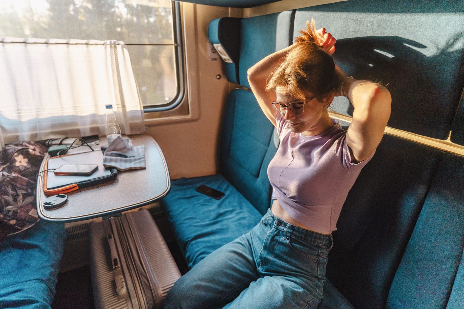 Young woman tying up her hair in a train sleeper compartment, suitcase and tablet near the window