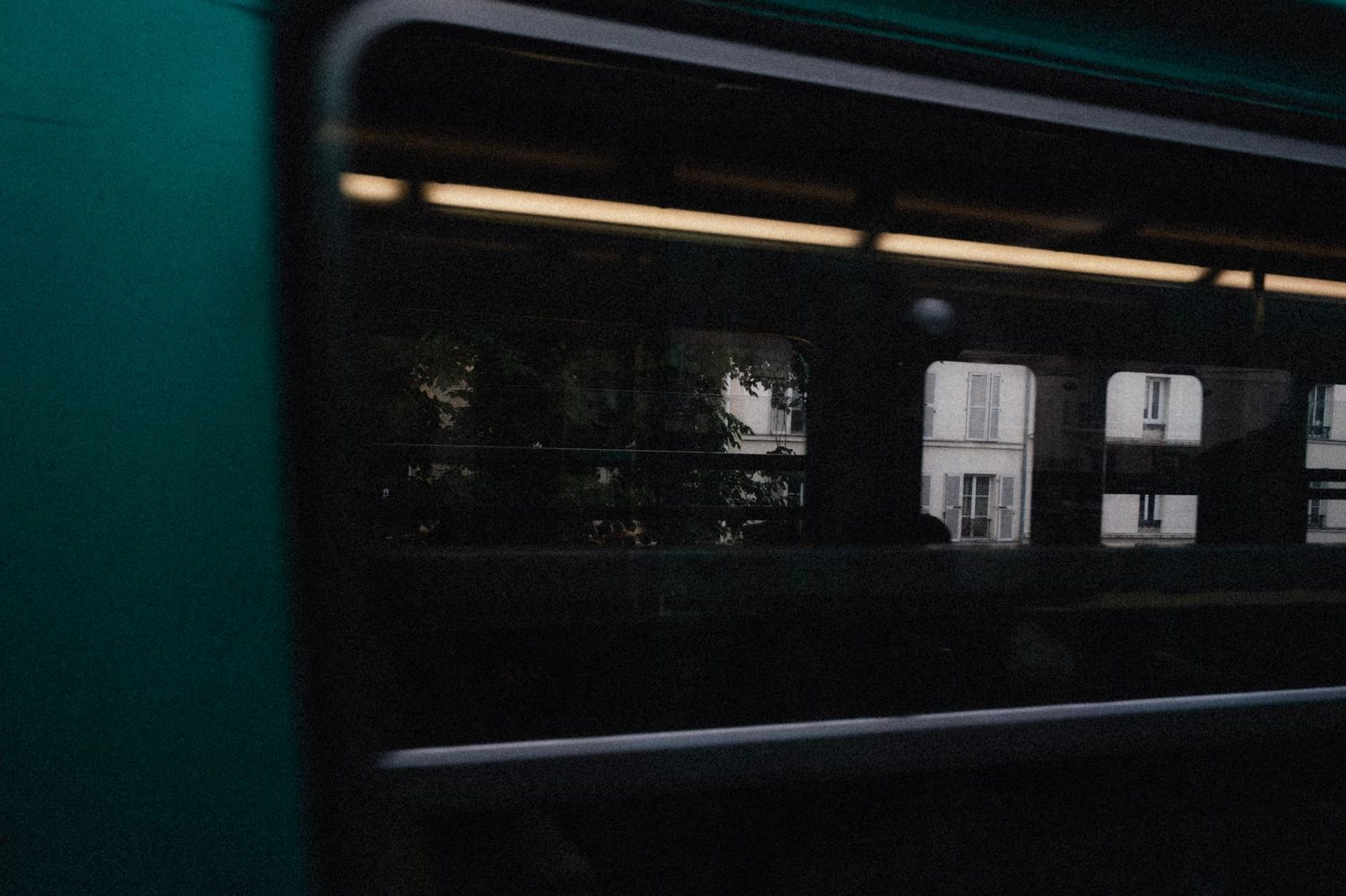 Fenêtre d'une voiture de train verte le soir, reflets d'arbres et façades d'immeubles éclairées