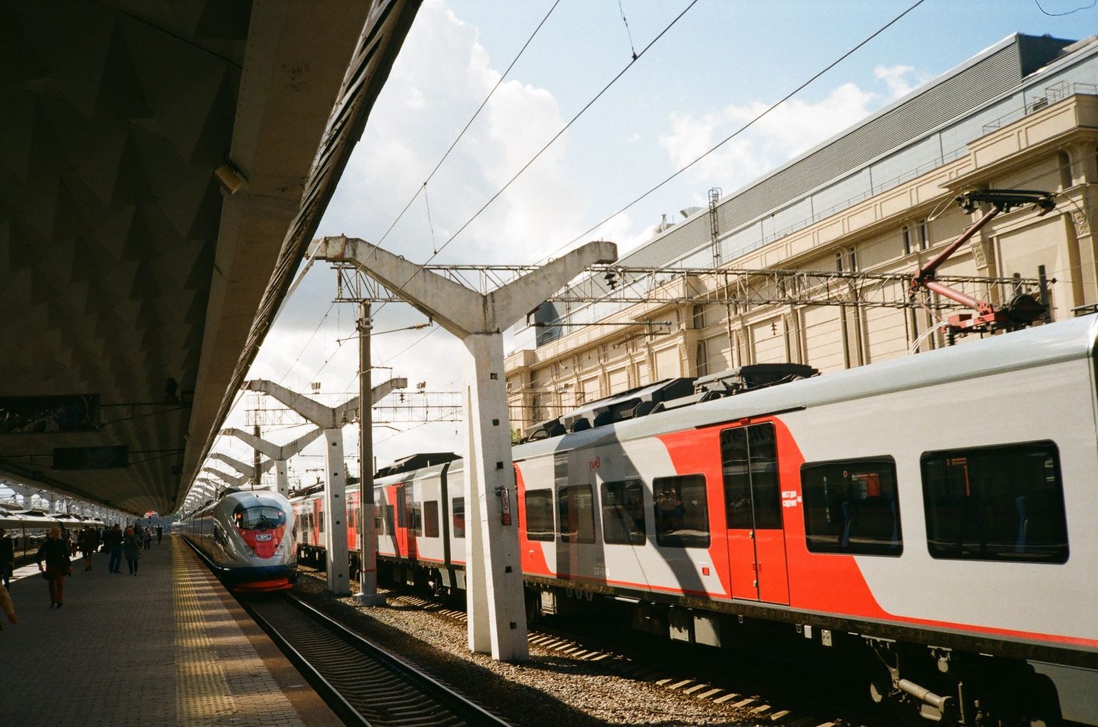 Trains at the platform beneath overhead lines in a sunlit station, travellers walking along the platform