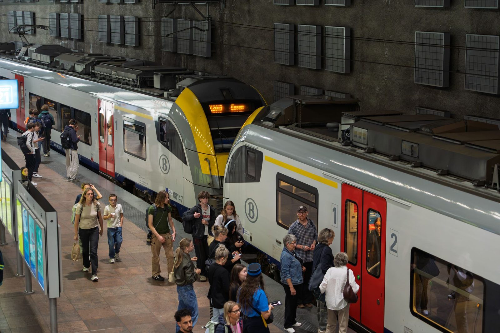 Reisende auf einem unterirdischen Bahnsteig zwischen zwei weiß-gelben belgischen Nahverkehrszügen