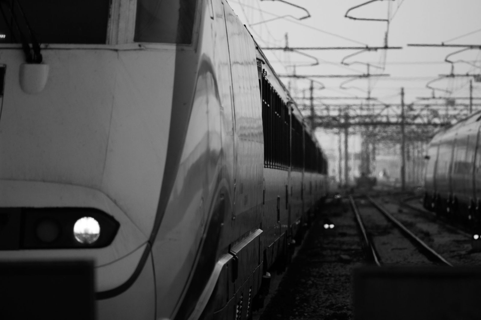 Black-and-white photo of a train at the platform alongside the tracks, under a station's overhead wires