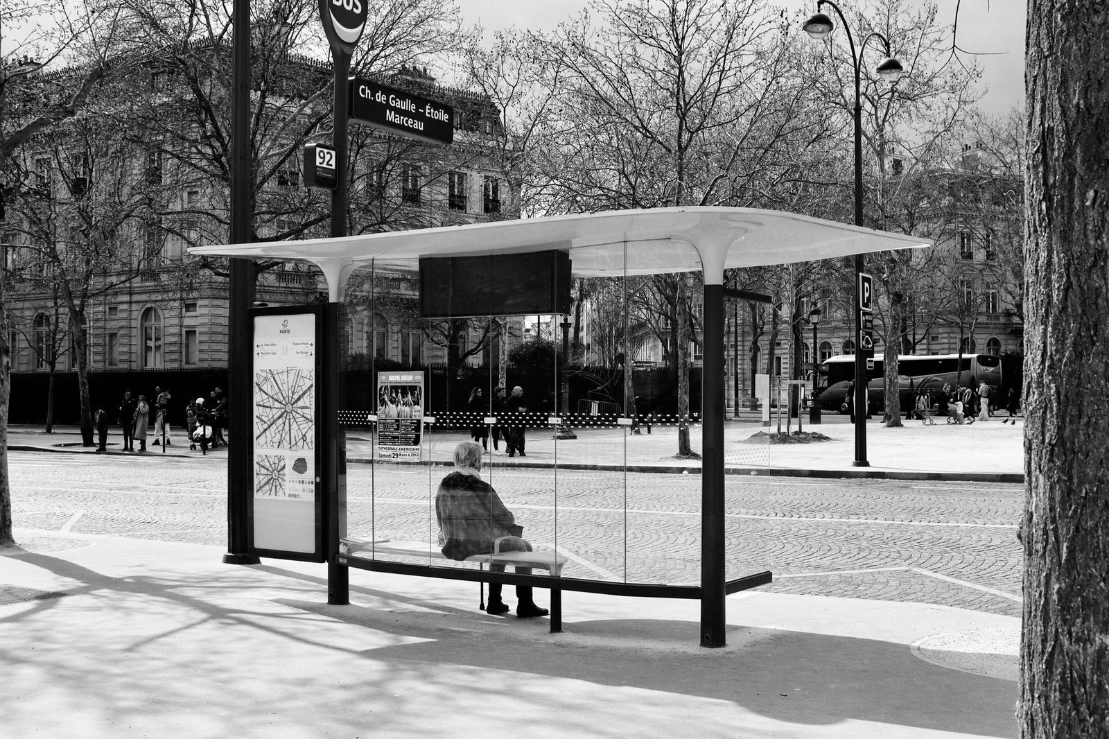 Glass bus shelter in Paris with a person sitting on the bench, buses and trees in the background, black-and-white photo