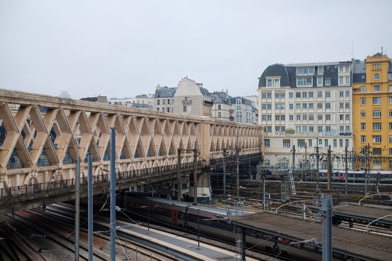 Faisceau de voies ferrées et train à l'approche d'une gare parisienne, sous un pont métallique et des immeubles haussmanniens