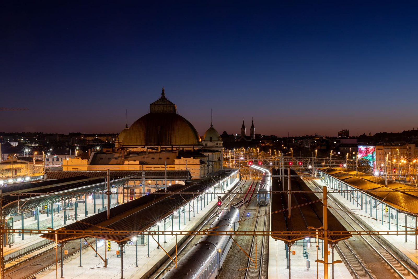 Estación de tren iluminada al atardecer, trenes en el andén y vías bajo las catenarias