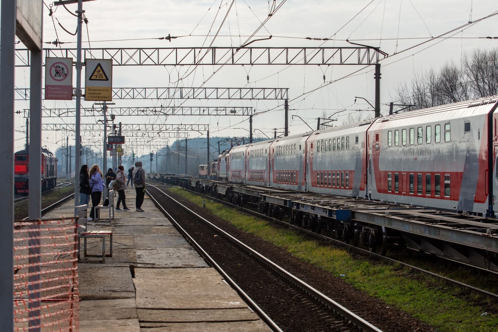 Reisende warten auf einem Bahnsteig, während ein Zug aus rot-weißen Wagen auf den Gleisen vorbeifährt