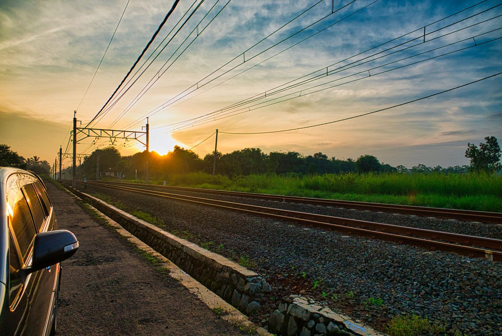 Electrified railway tracks and overhead lines at sunset, a car parked alongside the ballast