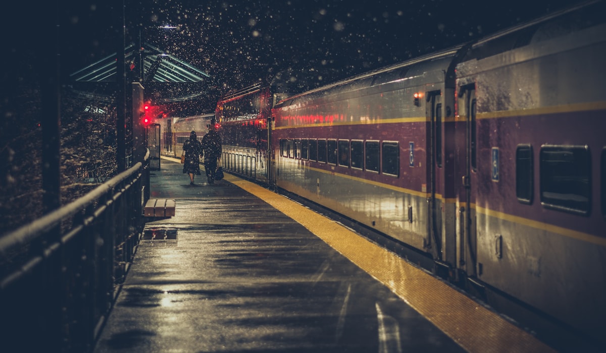 Un treno notturno fermo su una banchina europea innevata, con due passeggeri che camminano verso di esso con i bagagli