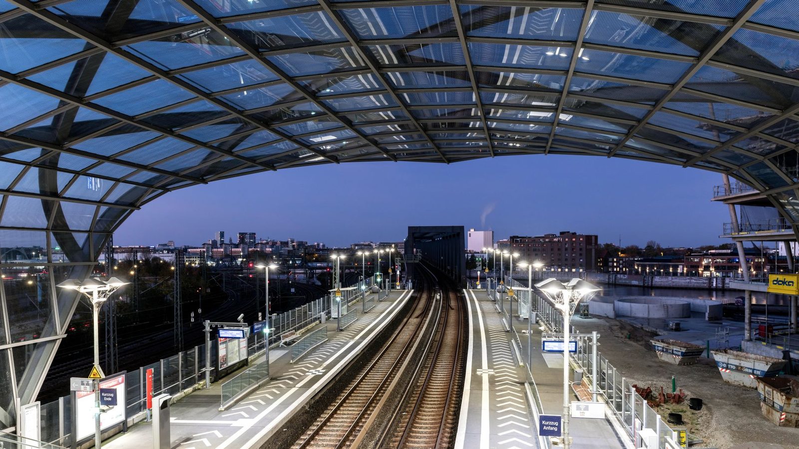 Platforms and tracks under the glass roof of an urban station lit at dusk