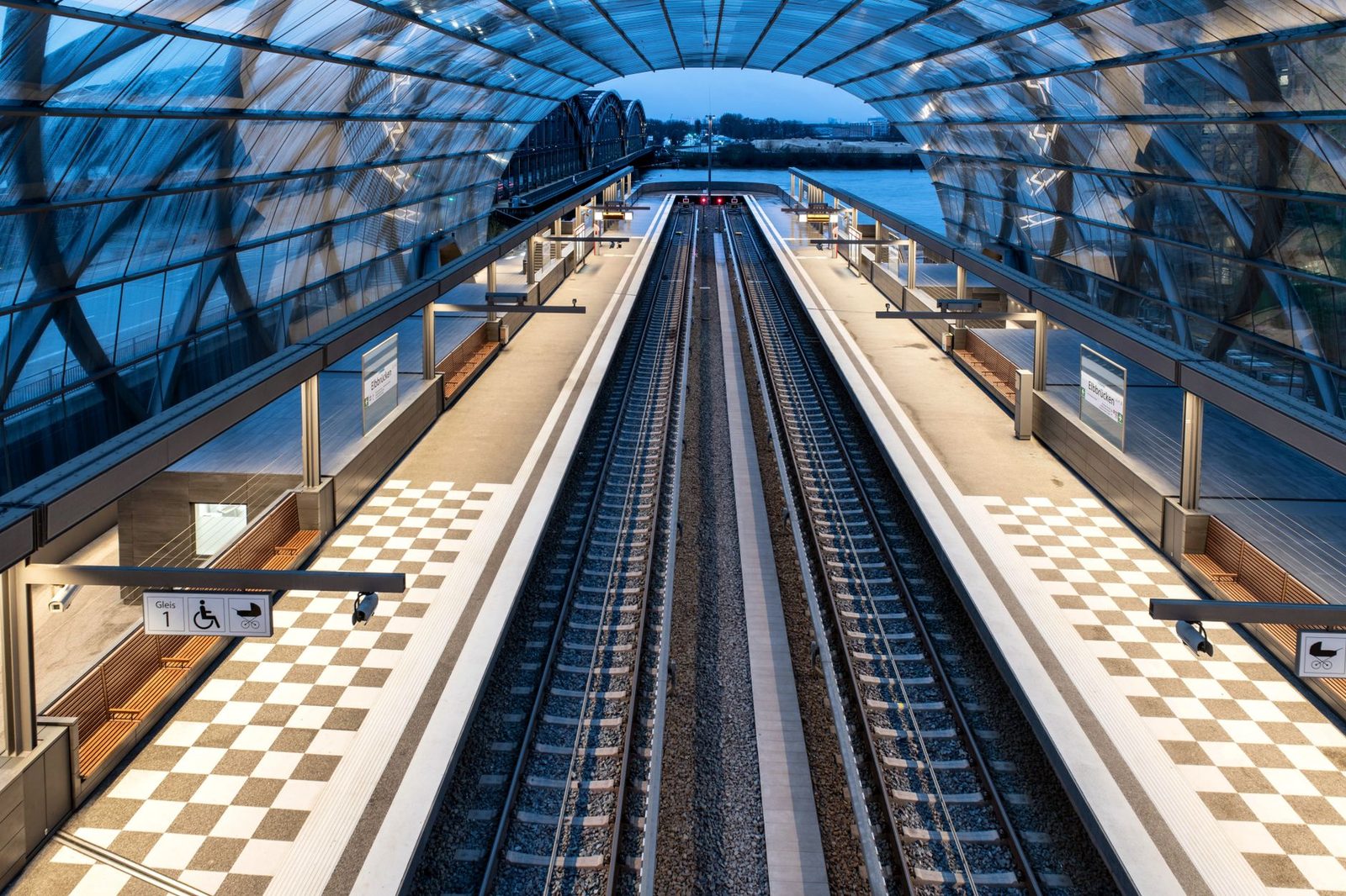 Deserted railway station platform under a modern glass canopy, with two tracks and a train in the distance at dusk