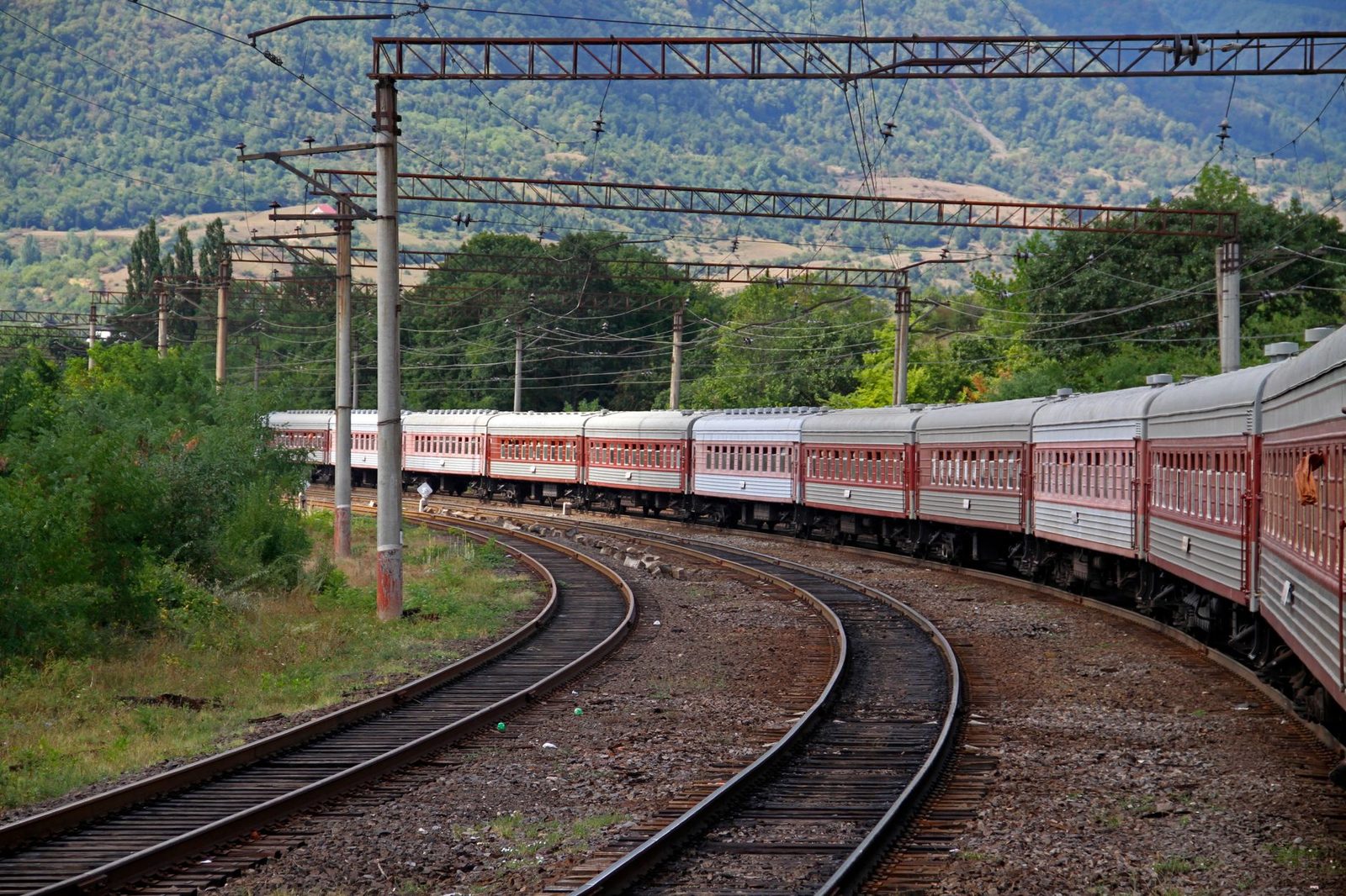 Tren de coches de viajeros rojos y blancos en una vía curva electrificada, colinas arboladas al fondo