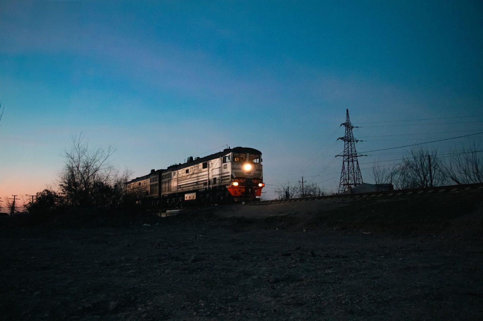 Von einer Diesellokomotive gezogener Zug mit eingeschalteten Scheinwerfern in der Dämmerung neben einem Strommast
