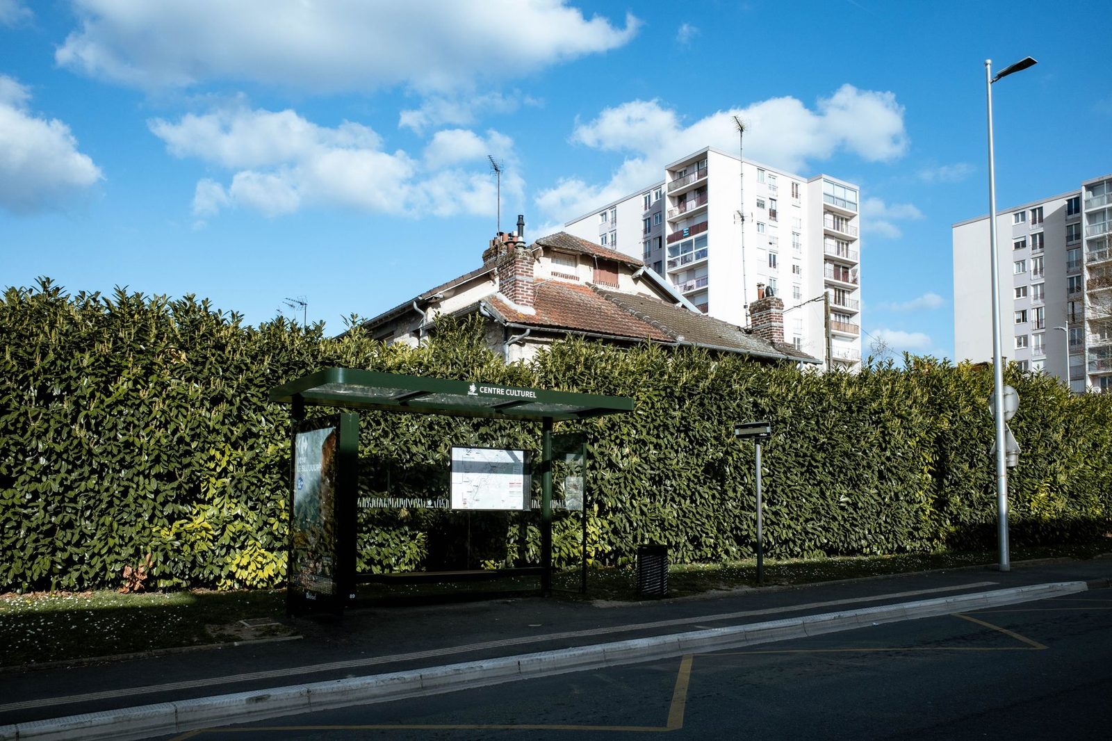 Abribus vert avec plan de ligne au bord d'une route, devant une haie et des immeubles