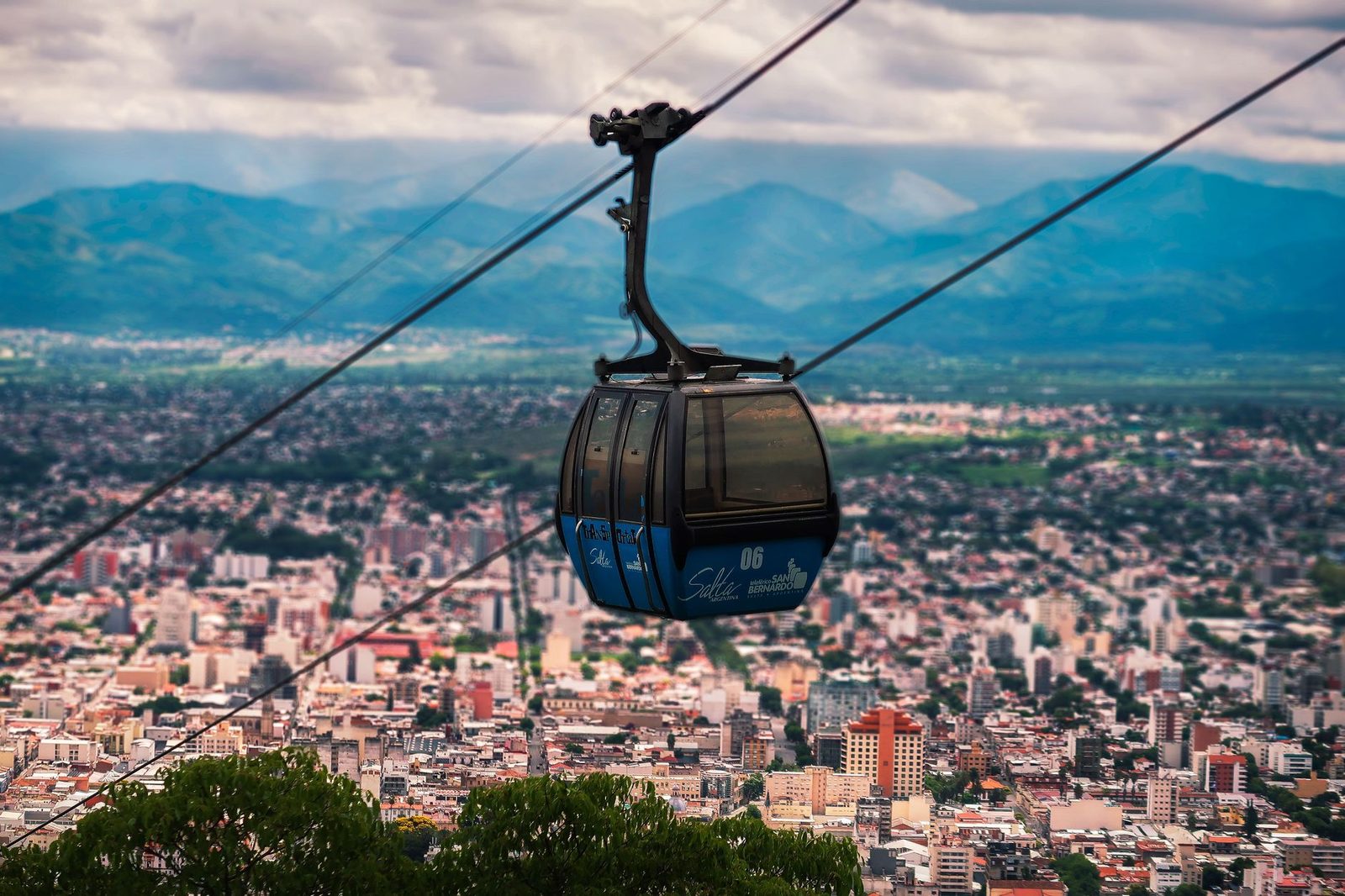 Cabine de téléphérique urbain bleue survolant une ville, montagnes et ciel nuageux en arrière-plan