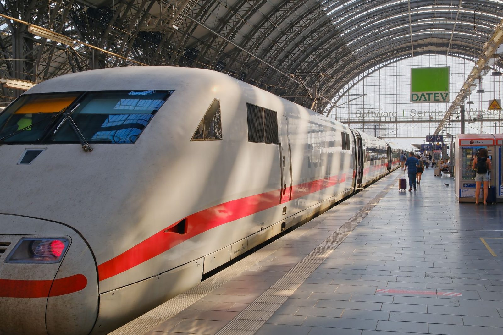 Weißer ICE-Hochgeschwindigkeitszug am Bahnsteig unter der Bahnhofshalle, Reisende mit Koffern auf dem Bahnsteig