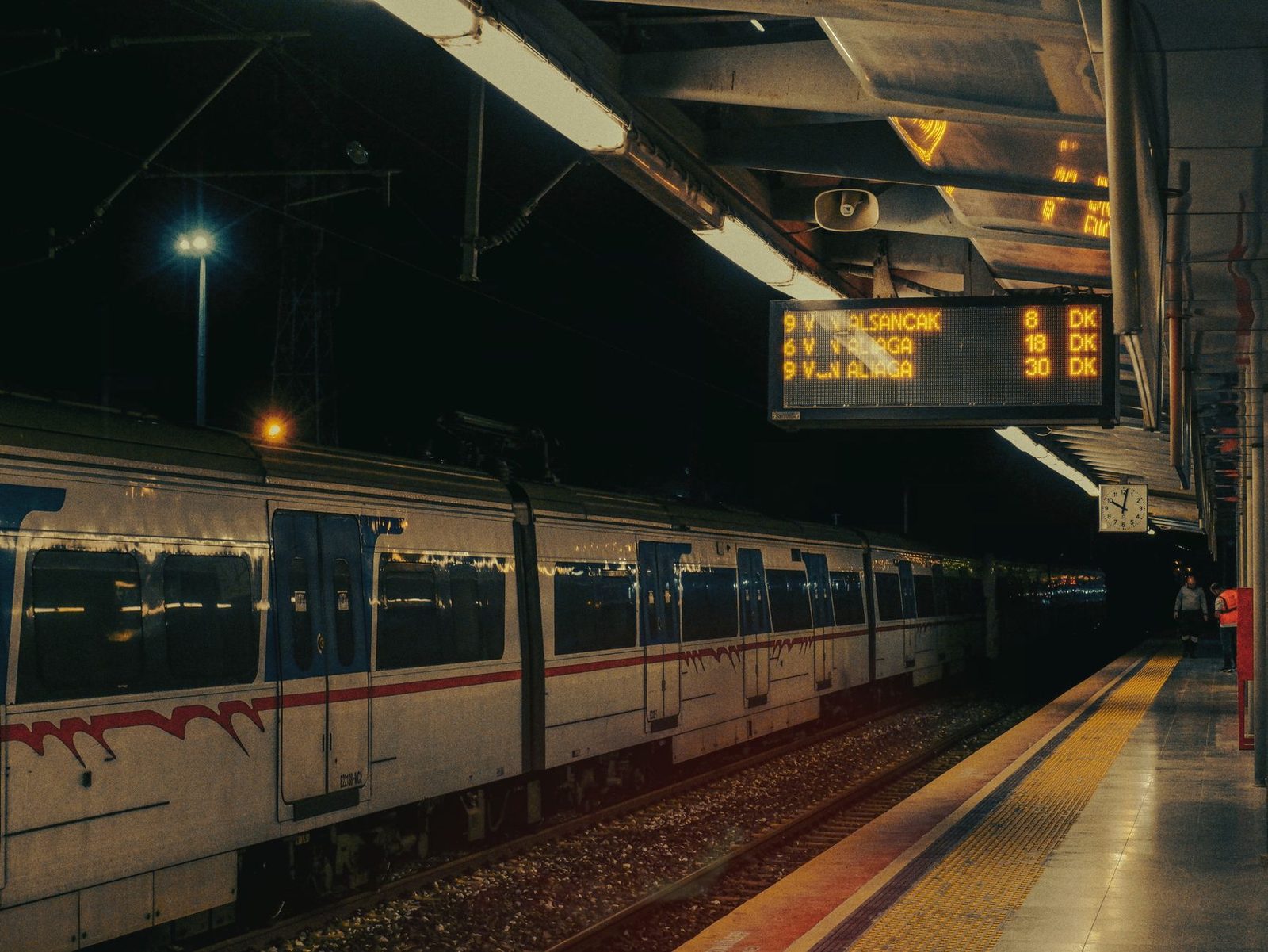 Train at the platform in a station at night, illuminated departure board and clock beneath the lit canopy