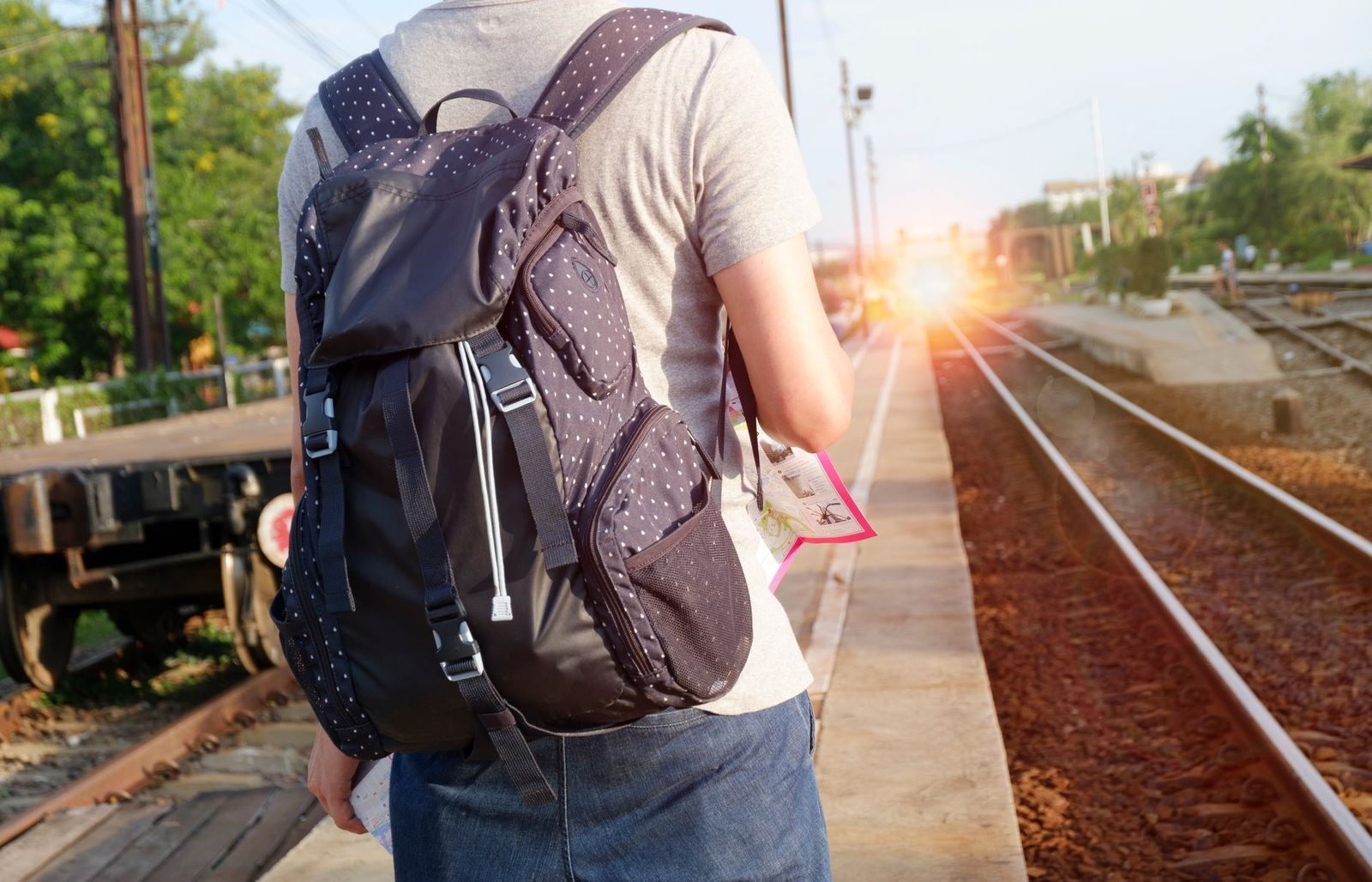 Traveller seen from behind with a backpack and ticket waiting on a railway platform at sunset